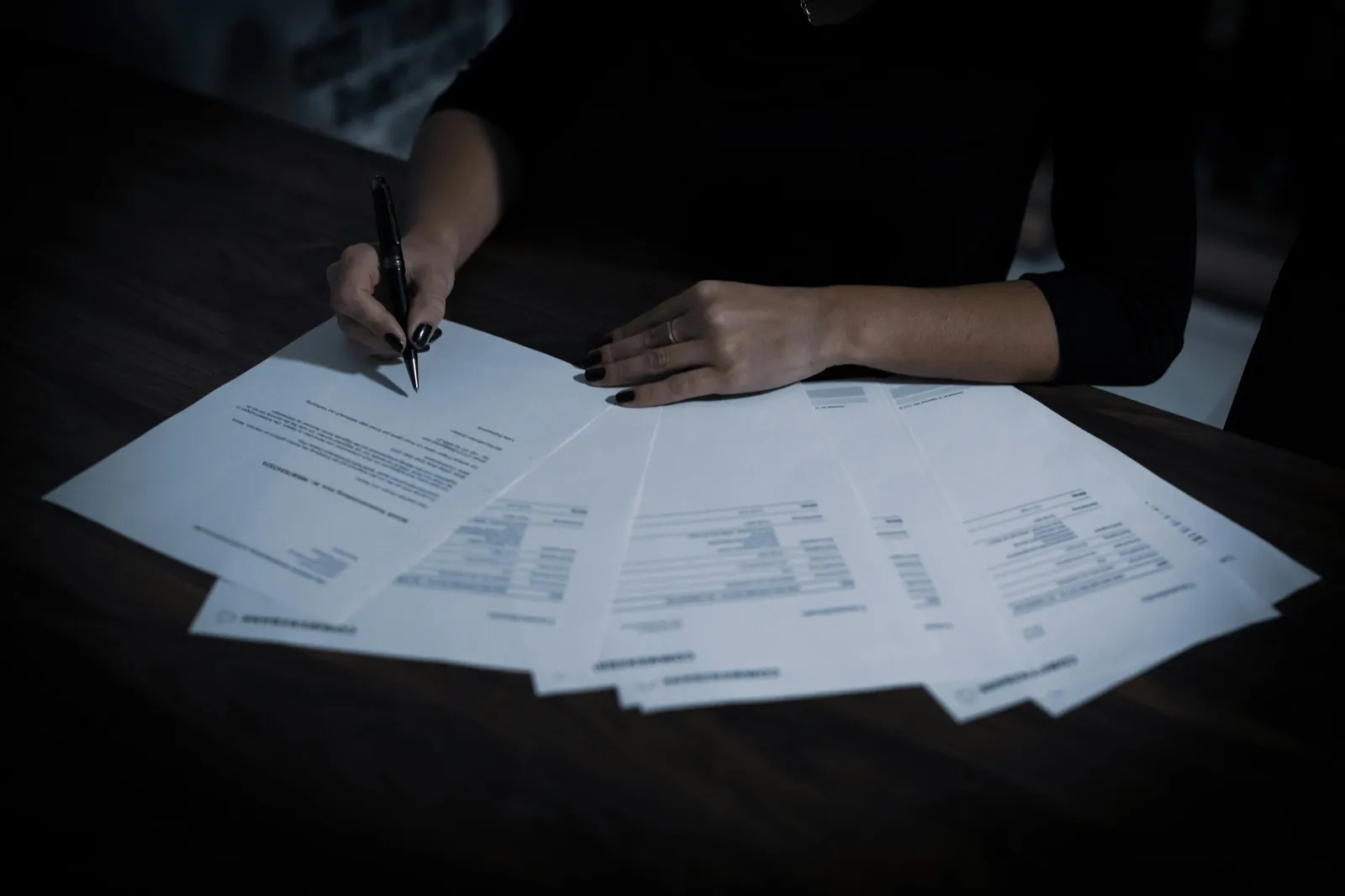 A person’s hands over fanned-out printed pages at a dark table, pen in hand