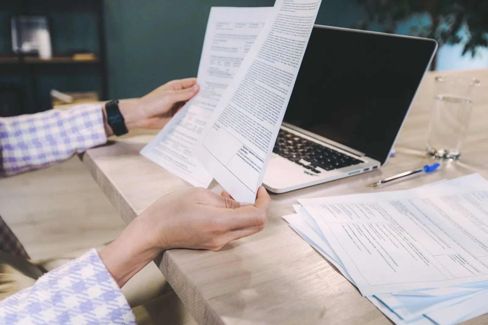 Two people’s hands holding printed pages beside an open laptop on a light wooden desk