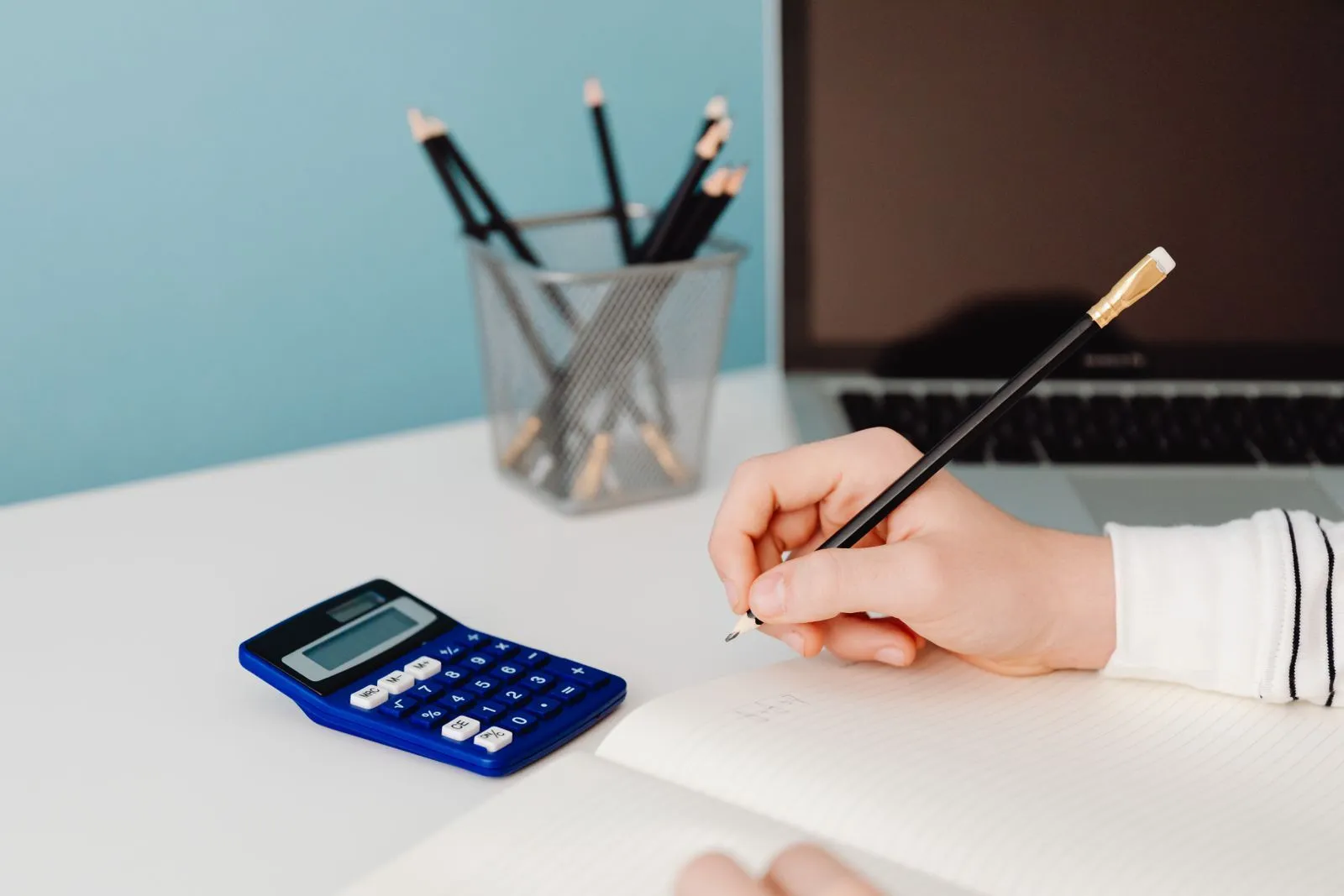 A hand writing in an open notebook beside a blue calculator and a pencil pot on a white desk