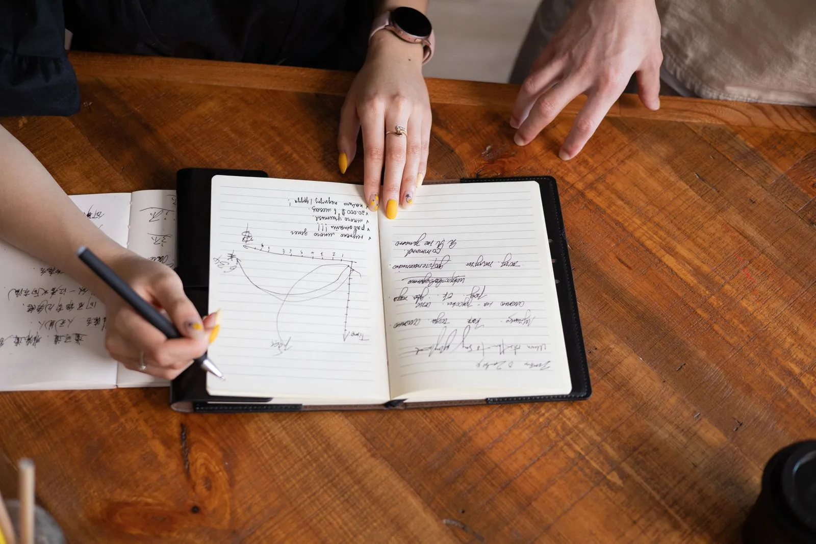 Overhead view of two people’s hands over an open notebook with a handwritten sketch, one writing with a pen
