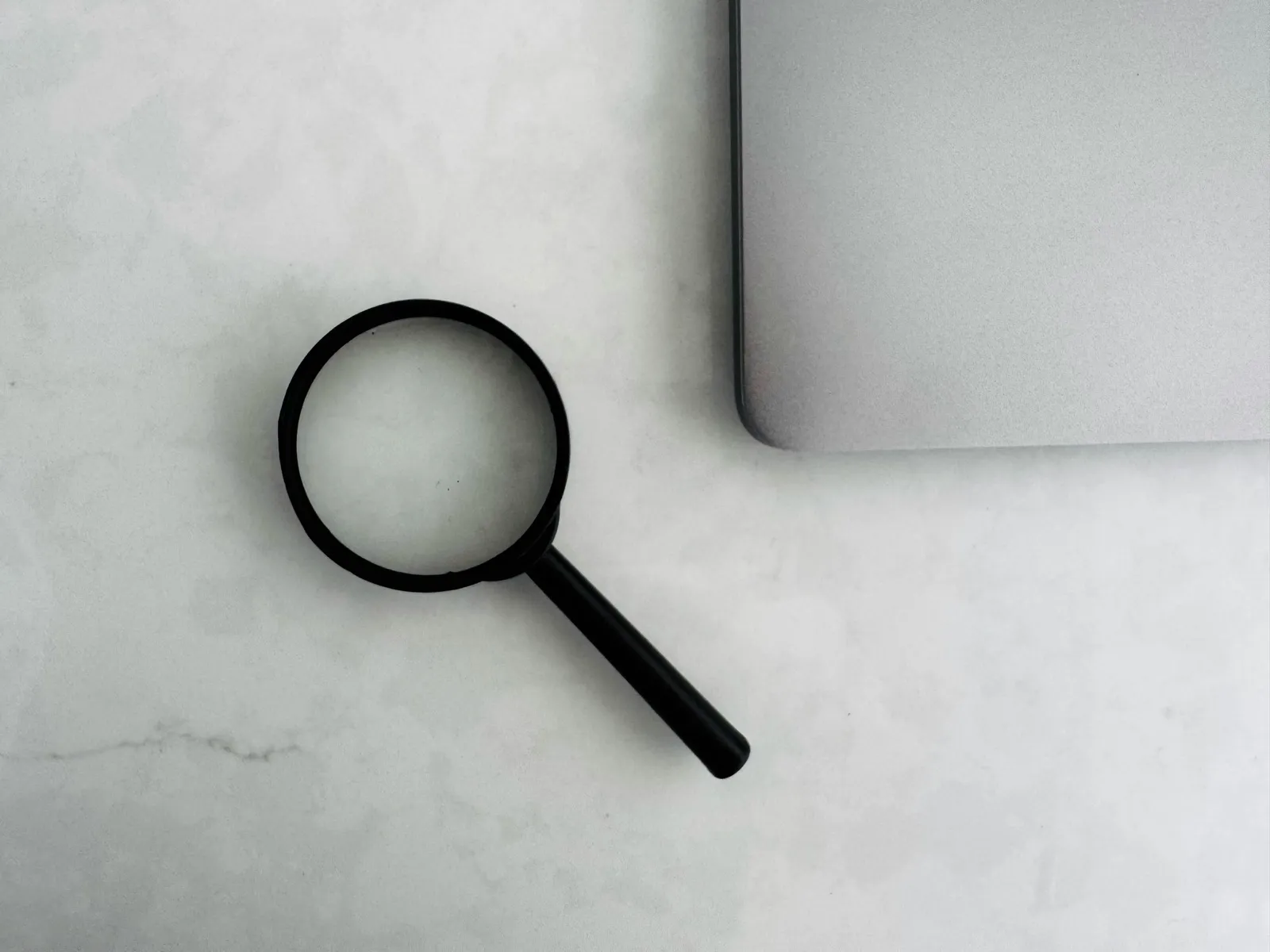 A black magnifying glass on a white marble surface beside the corner of a laptop